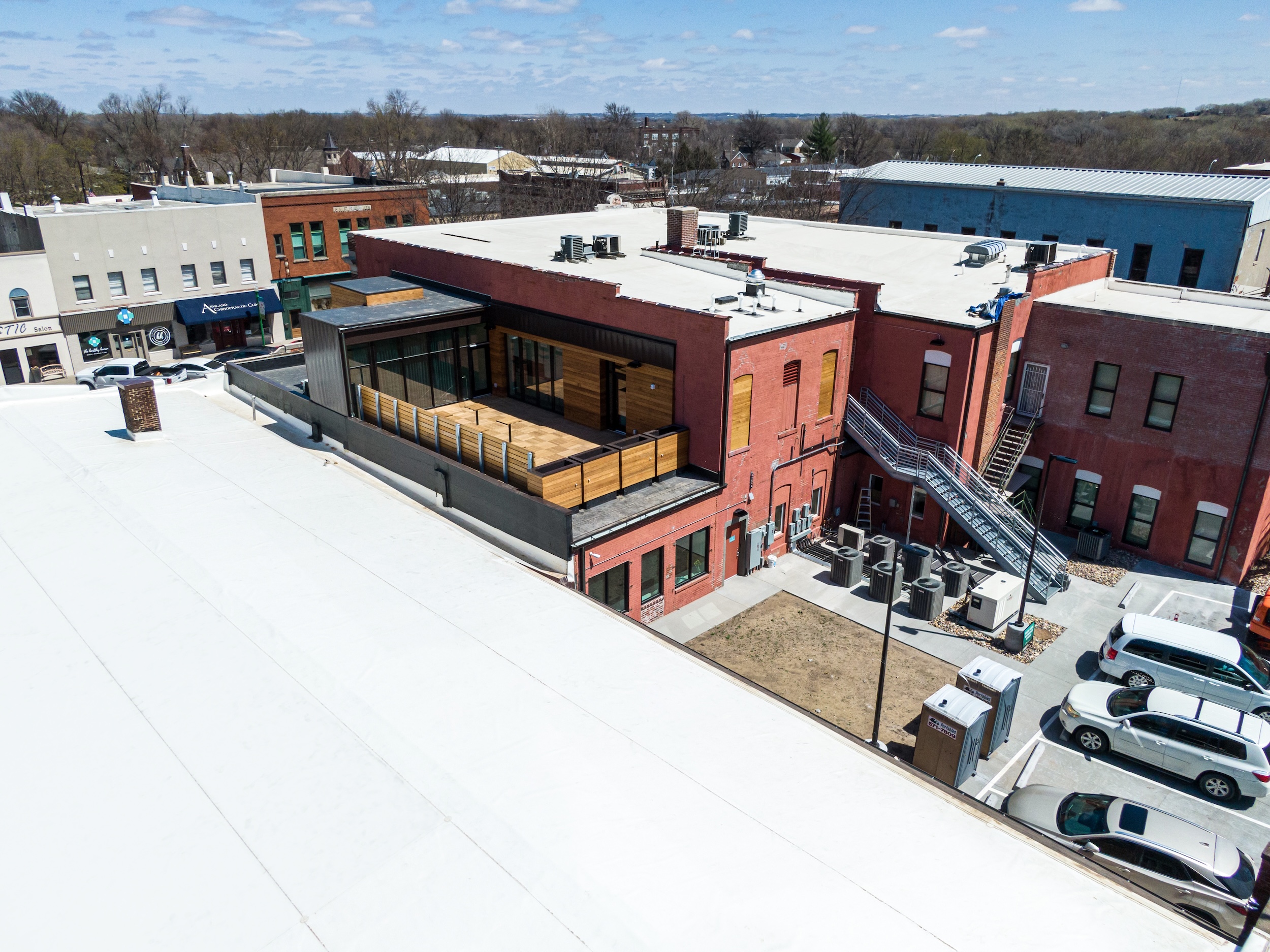 Aerial view of a renovated commercial building with a white GAF EverGuard TPO flat roof. The red brick structure features a modern rooftop deck enclosed with horizontal wood paneling and metal railings. Adjacent buildings and a small parking lot with vehicles are visible, with a metal staircase providing building access. The setting is a small downtown area on a partly cloudy day.