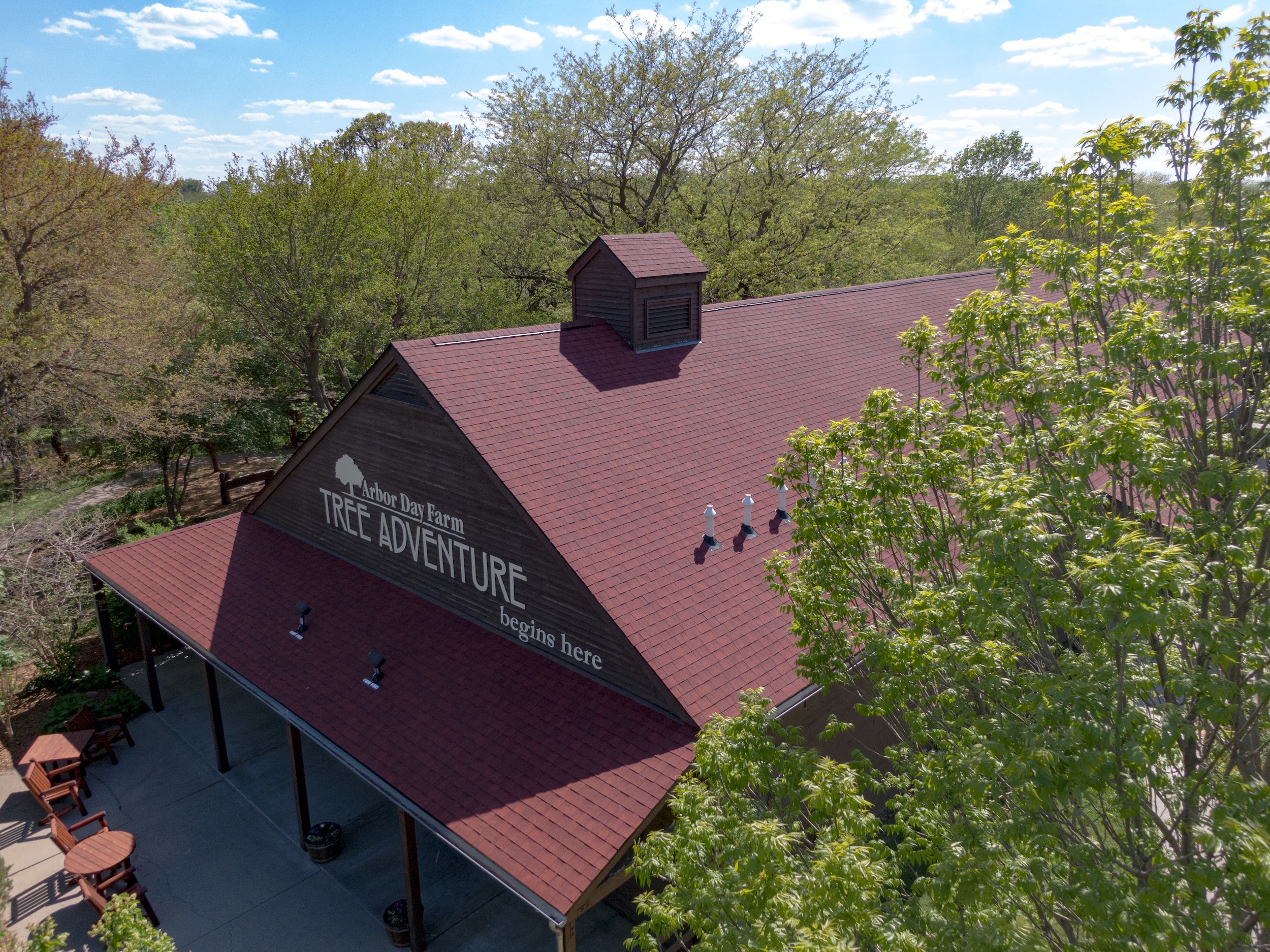 An aerial close-up view of the front of the Arbor Day Farm Tree Adventure building, featuring a bold GAF Timberline HDZ roof in Patriot Red, installed by Roof Rite. The rich red shingles contrast with the dark wood-toned gable and adjacent trees. White plumbing pipes and a central cupola add architectural detail. The covered entrance patio includes outdoor seating and signage inviting visitors to explore.