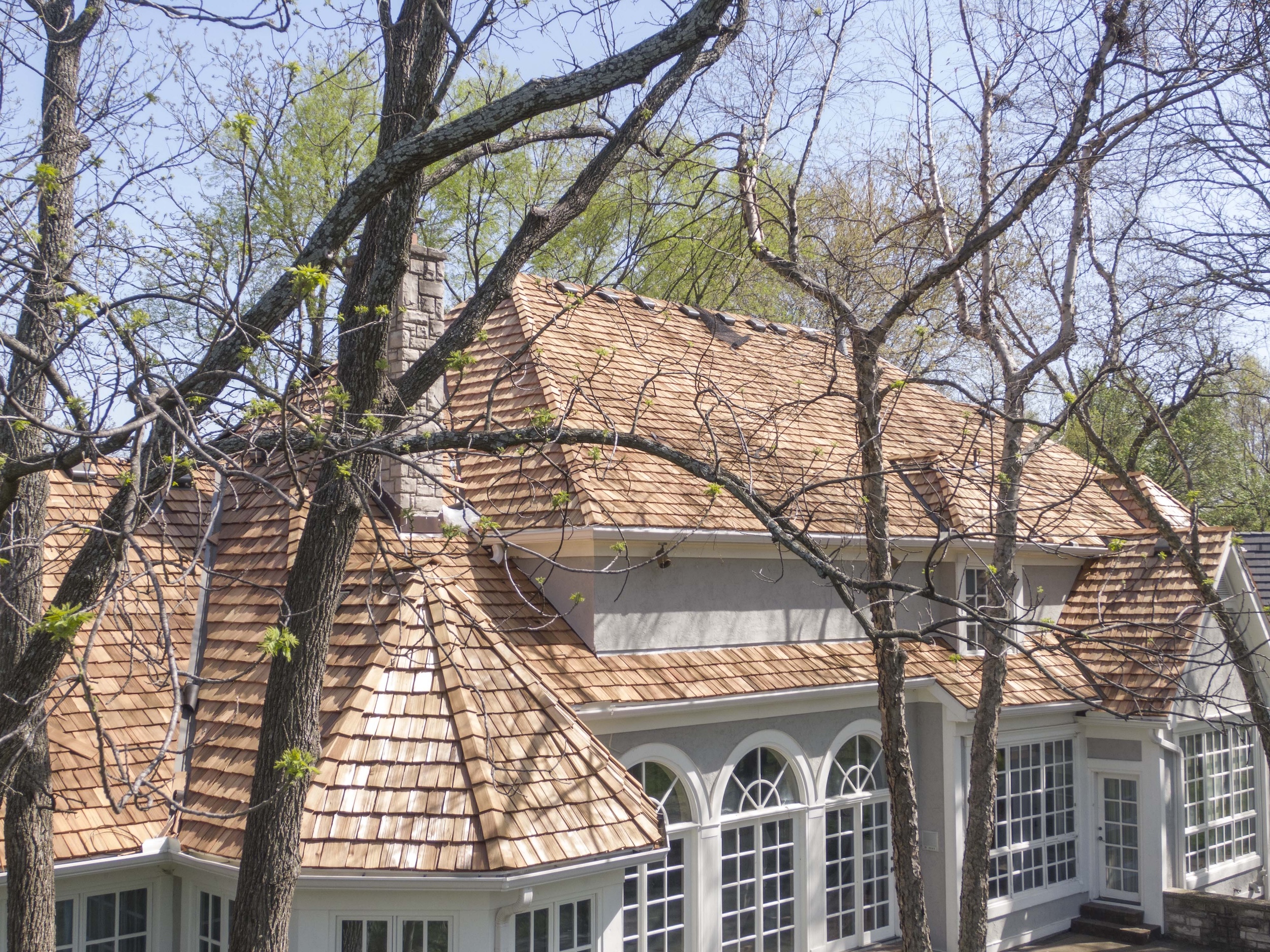 Elegant residential home with freshly installed natural cedar shake roofing installed by Roof Rite, in an established, wooded area. The roofline and victorian turret complement the home's arched windows, gray stucco walls, and stone chimney. Surrounded by tall trees and landscaped greenery, the cedar shakes add a warm, classic aesthetic to the property.