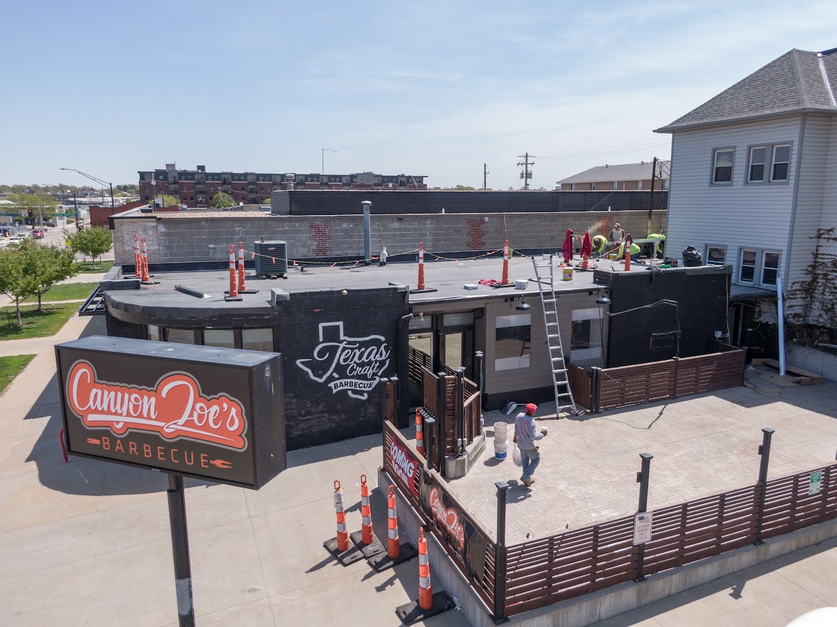 Aerial angled view of Canyon Joe's Barbecue during Roof Rites, Carlisle EPDM flat roof installation. Crews are working on the roof above the black exterior, with safety cones and tools visible across the surface. The project sits in a mixed-use neighborhood with adjacent residential and commercial buildings.