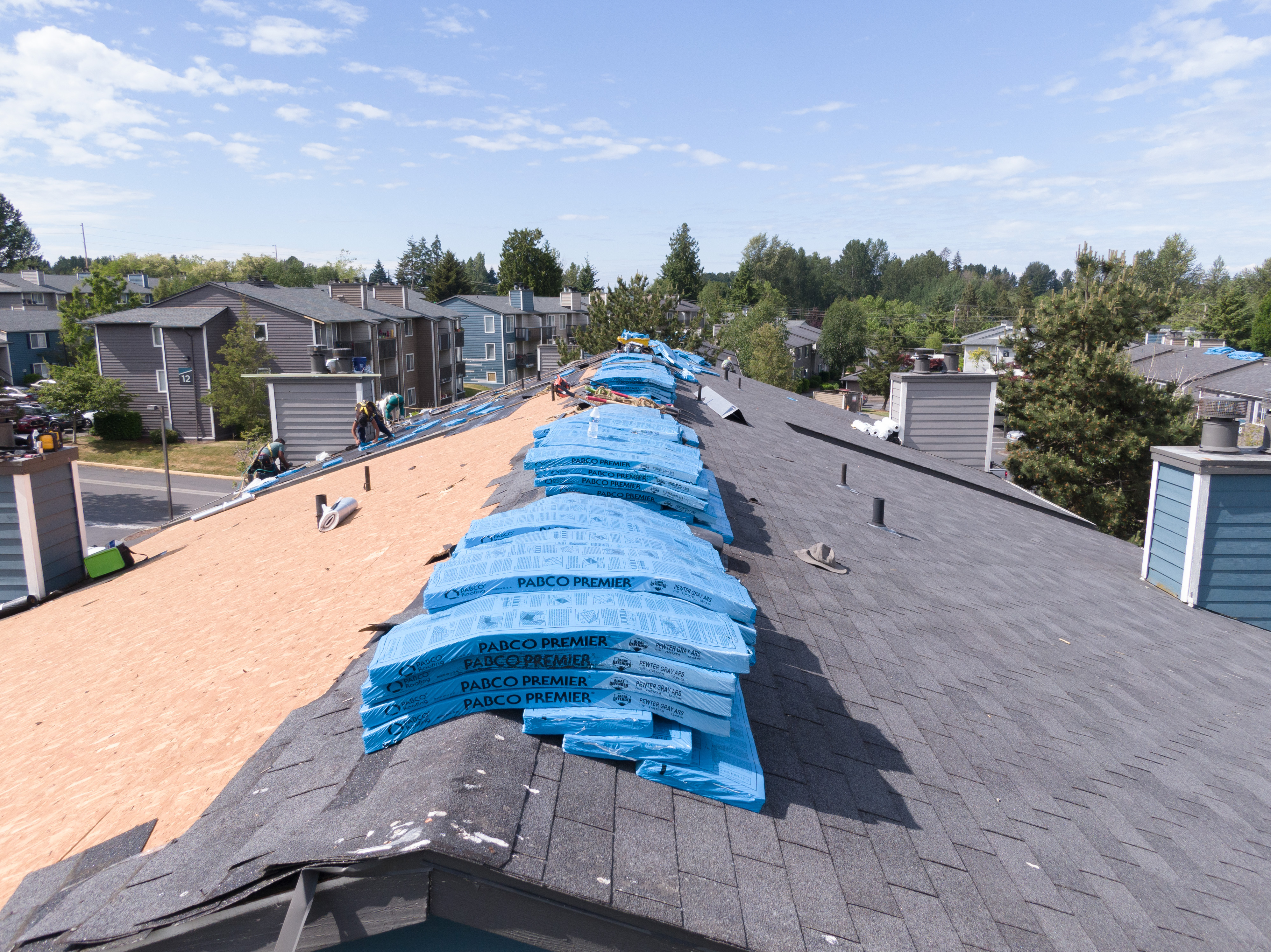 A partially-finished roof figures prominently in the image. In the background, three workers in personal protective equipment are fastening starter shingles to the underlayment. The ridge of the roof is being used to store shingles, providing easy access to the roofing workers as they diligently affix the building's new roof to its decking.