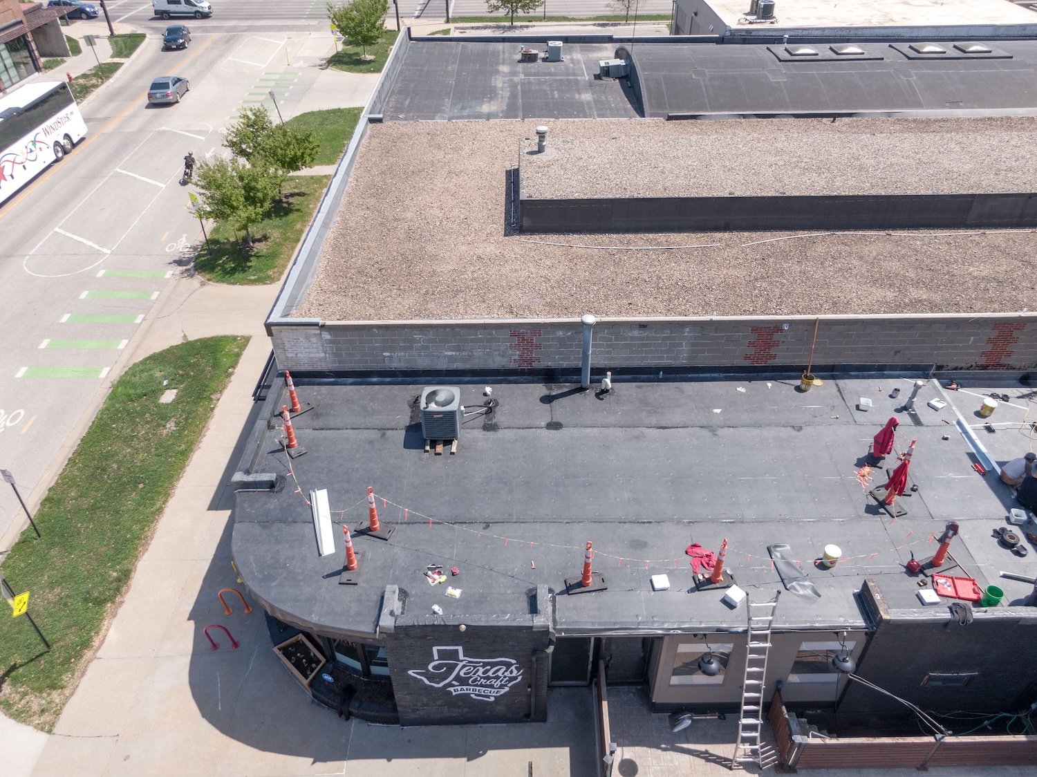 Aerial view of a restaurant building with Roof Rite in the middle of a flat roof replacement. The Carlisle EPDM Membrane is partially completed with tools, safety cones, and materials visible across the surface. The building labeled 'Texas Craft Barbecue' sits on a street corner with a rounded front entryway adjacent bike lanes, and light traffic.