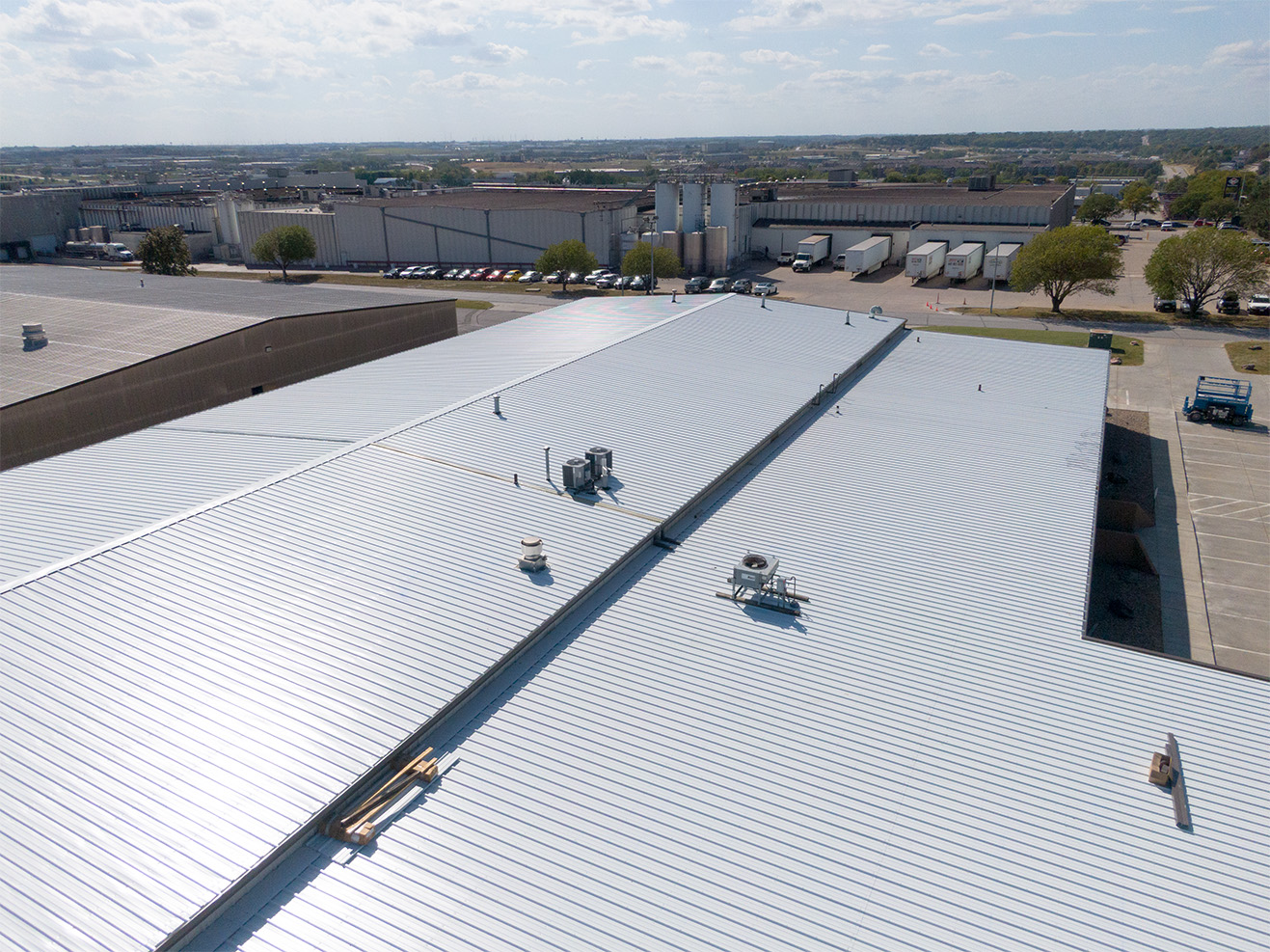 Aerial view of a large industrial facility with a new standing seam metal roof installed by Roof Rite. The clean, reflective surface spans multiple connected structures and includes various rooftop units and penetrations. The photo showcases the scale and precision of the commercial roofing project, with clear blue skies and a mix of industrial buildings, parking lots, and delivery trucks visible in the background.