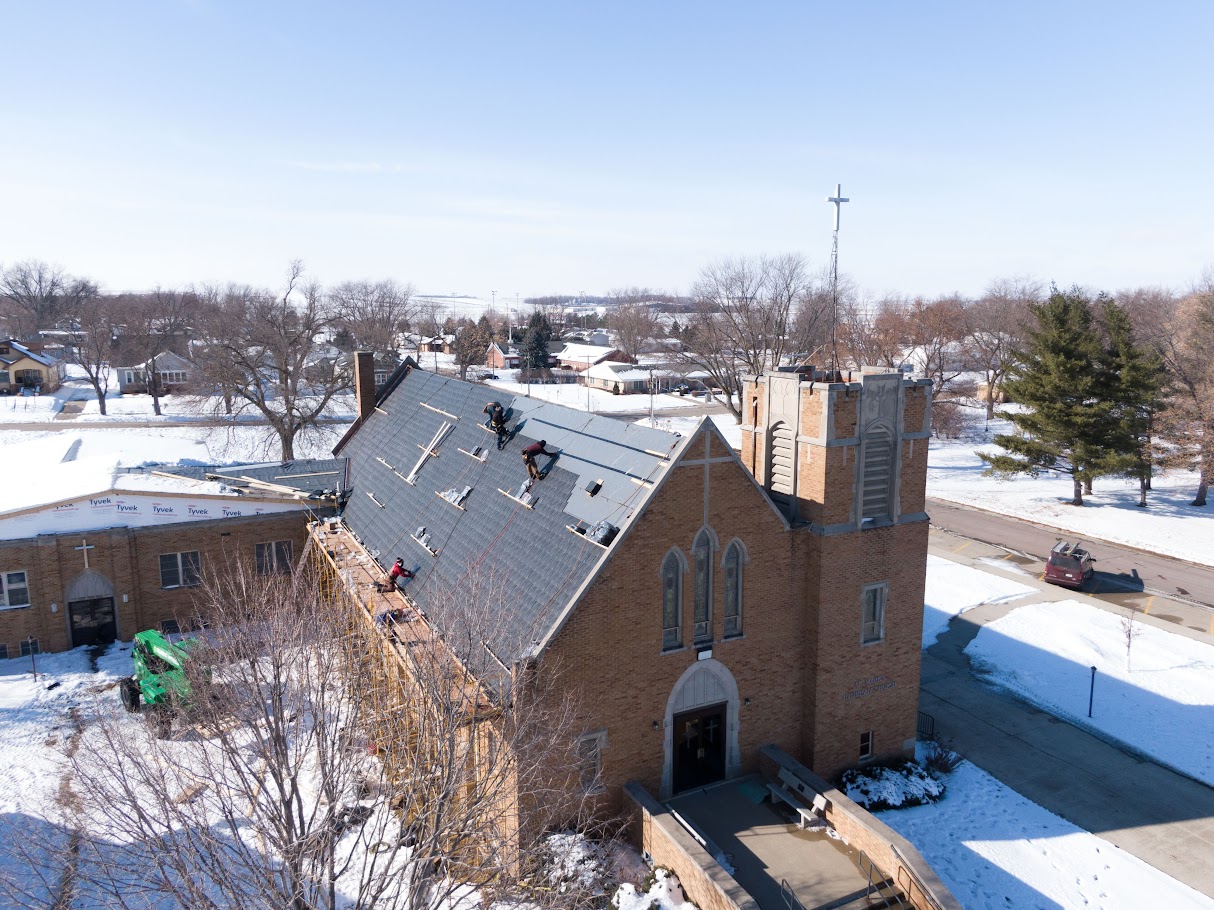 Aerial view of a church undergoing roof replacement by Roof Rite during winter. Workers are installing a new roofing system on the steep main sanctuary roof, with safety lines and materials visible. Snow blankets the surrounding ground and neighborhood, contrasting with the exposed roof decking. The brick church features gothic-style windows and a tower topped with a tall cross.