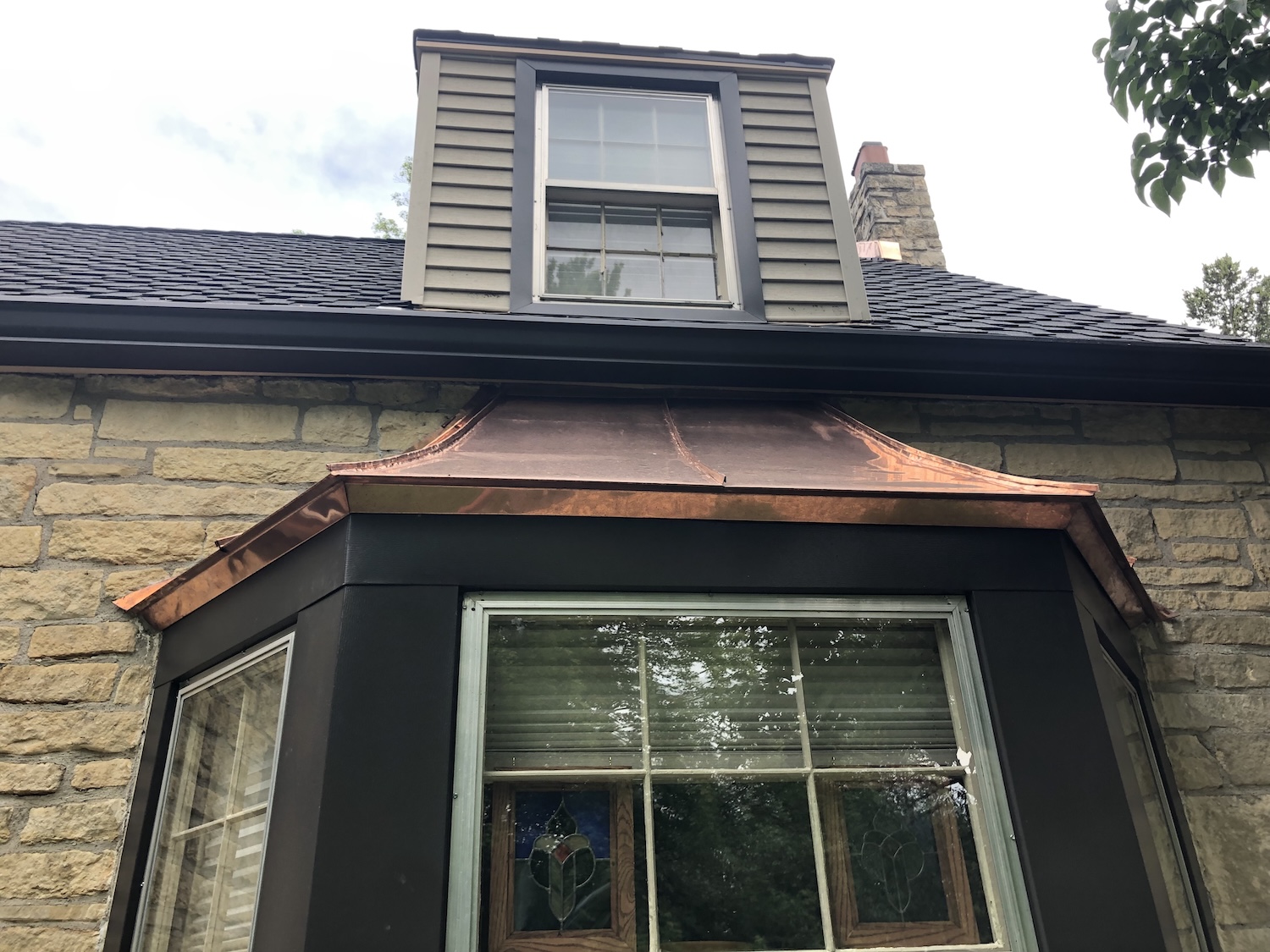 Close-up of a home's bay window, showing off Roof Rite's craftsmanship. The newly installed, patina-free copper pent roof contrasts with the dark metal window trim and stone exterior, adding an elegant architectural detail beneath a dormer window, and presidential shingle roof. The homeowner's stained glass projects hang framed, behind the window.