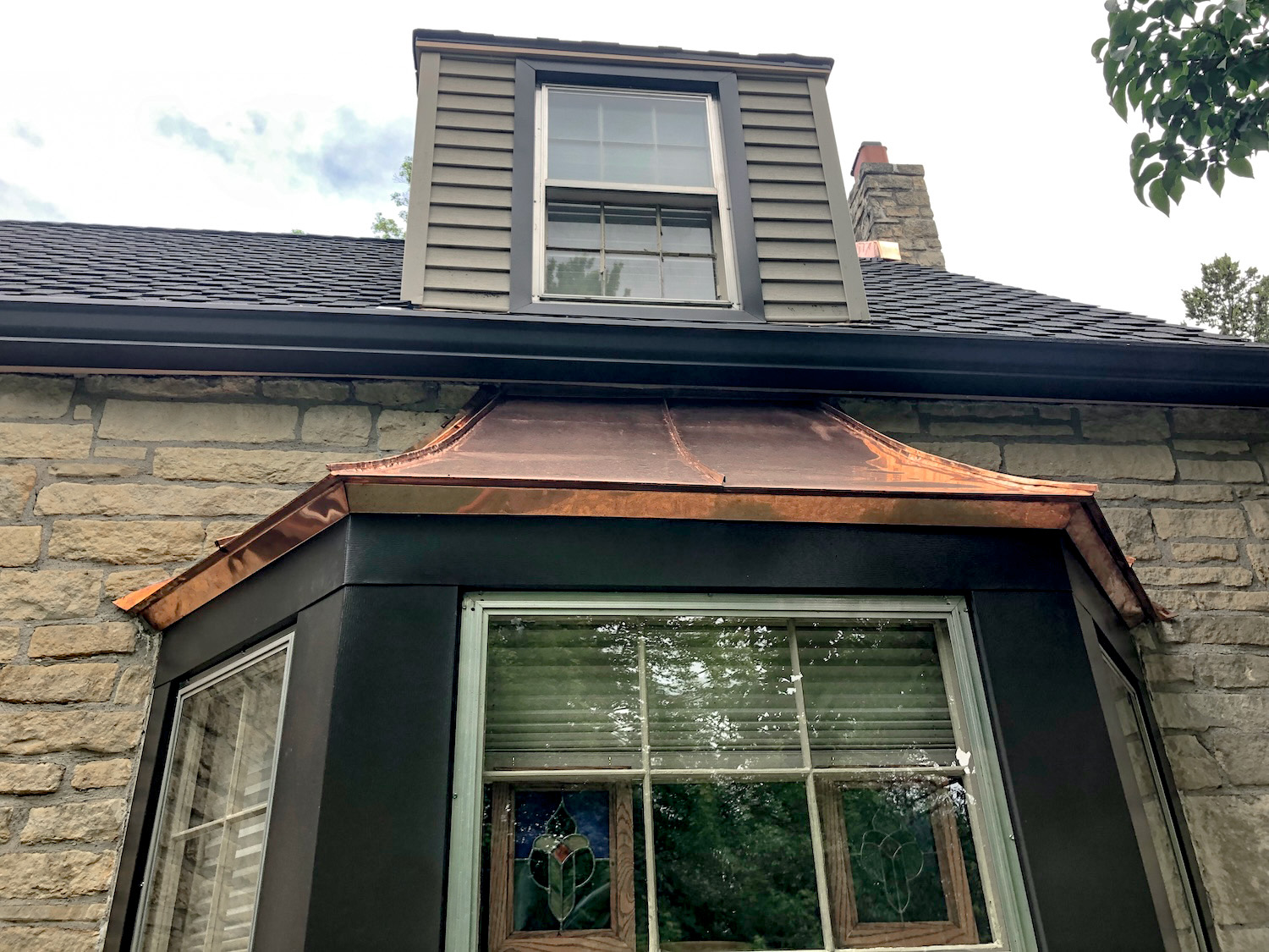 Close-up of a home's bay window, showing off Roof Rite's craftsmanship. The newly installed, patina-free copper pent roof contrasts with the dark metal window trim and stone exterior, adding an elegant architectural detail beneath a dormer window, and presidential shingle roof. The homeowner's stained glass projects hang framed, behind the window.