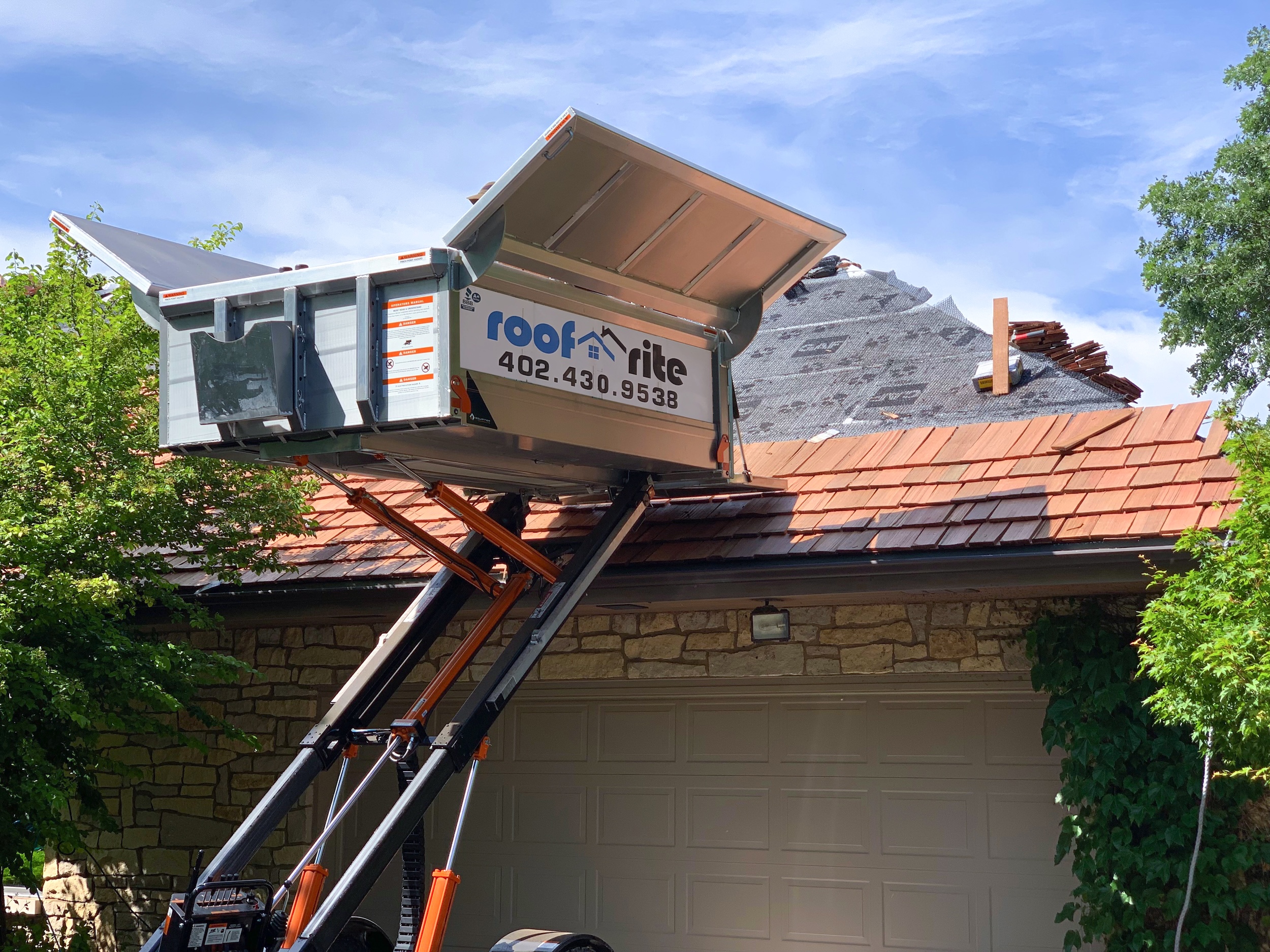 Roof Rite's Equipter RB5000 Self-propelled dump trailer raises it's bucket to the roofline of a stone house during a cedar shake replacement. The helps to manage debris during tear off, and safely load materials during replacement. The first few courses of shakes have already been installed, with the underlayment visible towards the ridge. Trees frame the garage below.