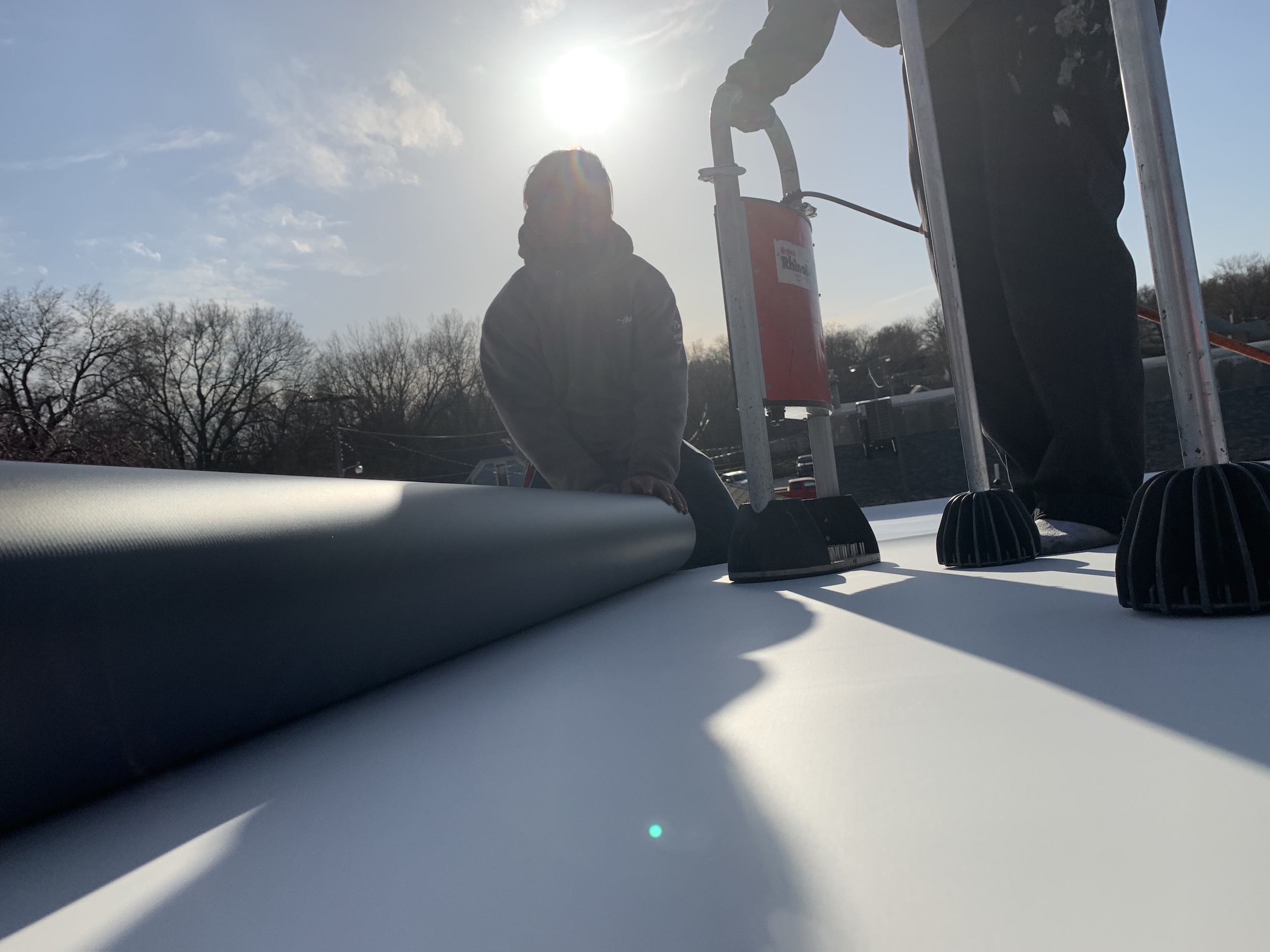 Close-up of a Roof Rite crew installing a white PVC roofing membrane on a flat roof. One worker unrolls the membrane while another operates a RhinoBond induction welder to secure the material. The sun shines brightly overhead, casting long shadows across the smooth, reflective surface. Leafless trees and rooftop HVAC units are visible in the background.