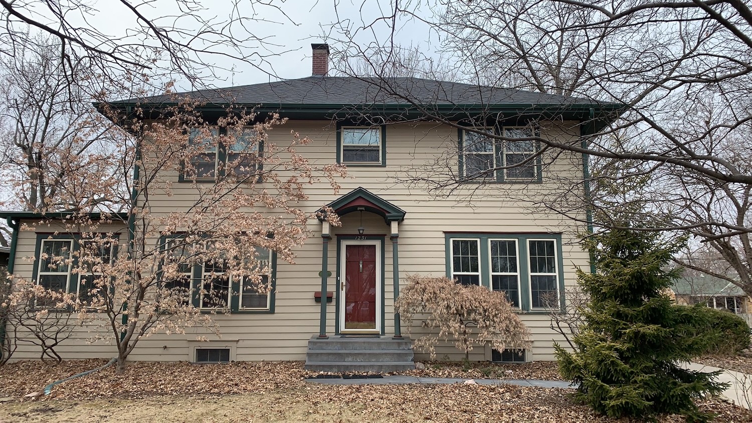 Front view of a two-story home with beige siding, green trim, and a deep red front door. The newly installed roof, completed by Roof Rite, features dark asphalt shingles that complement the house’s vintage charm. Bare trees frame the house, suggesting late fall or early winter, with dried leaves scattered across the lawn and walkway.