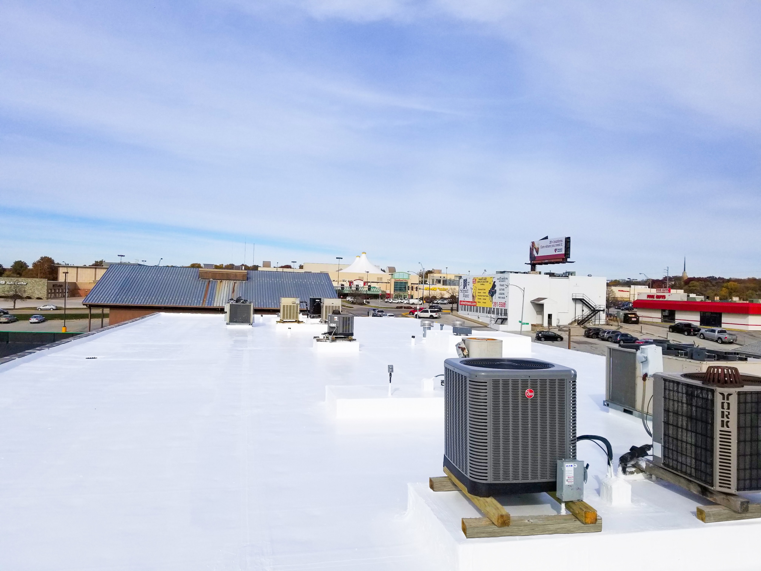 Flat commercial rooftop with a finished bright white elastomeric coating by Roof Rite. Several HVAC units and vents are installed. In the background, a commercial district with billboards, shopping centers, and parked vehicles is visible under a blue sky with soft cloud coverage.