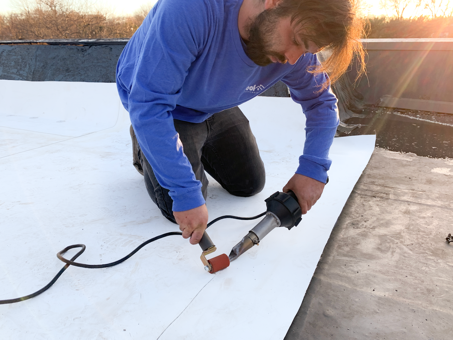 A Roof Rite team member uses a heat welder to seam white TPO roofing membrane on a flat roof at sunset. He kneels over the material with a hot-air gun in one hand and a silicone roller in the other, ensuring a secure bond.