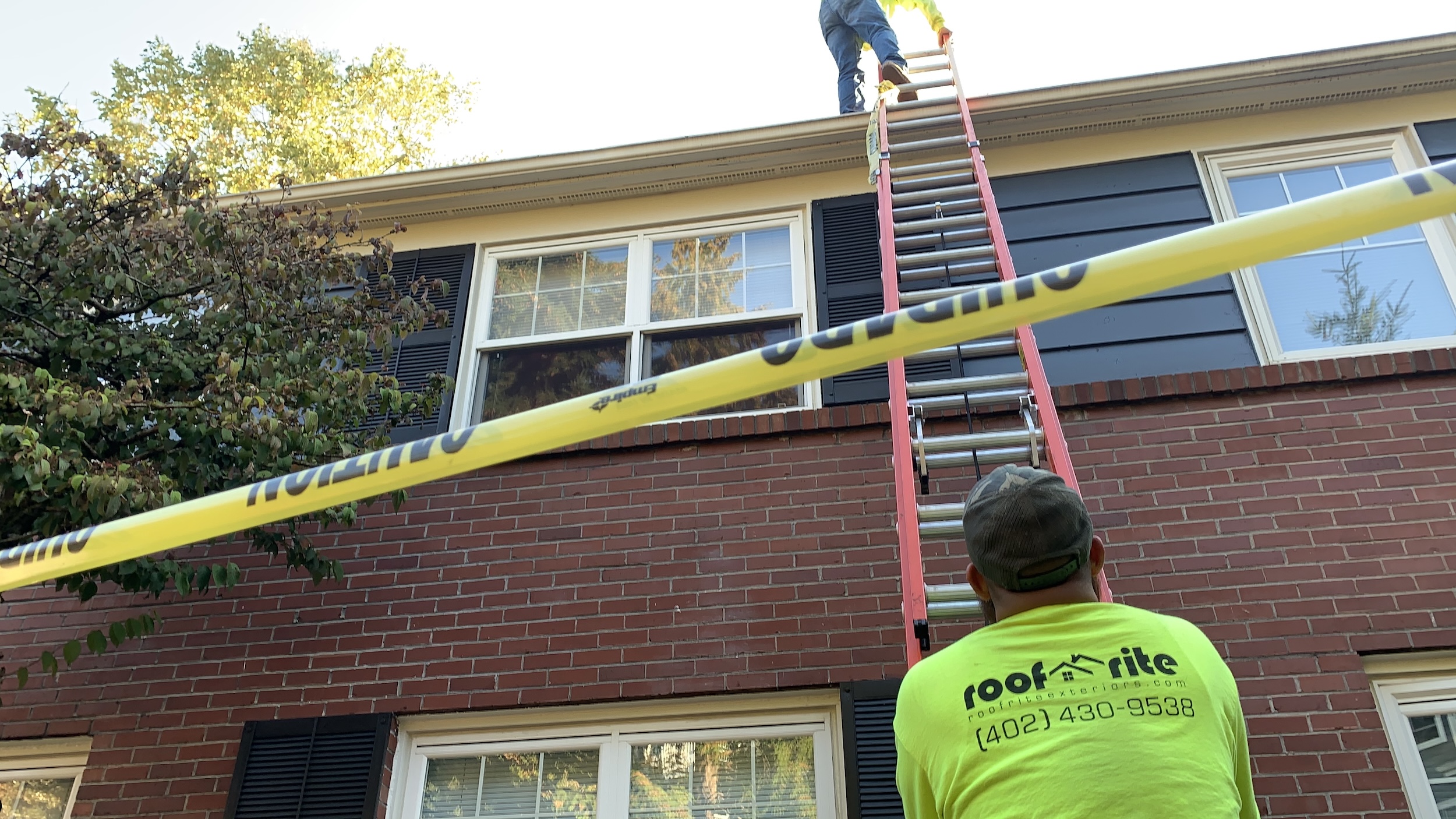 A Roof Rite team member keeps a ladder steady as his coworker ascends to the roof of a two-story brick building. Bright yellow caution tape stretches across the foreground. The crew is outfitted in high-visibility gear.